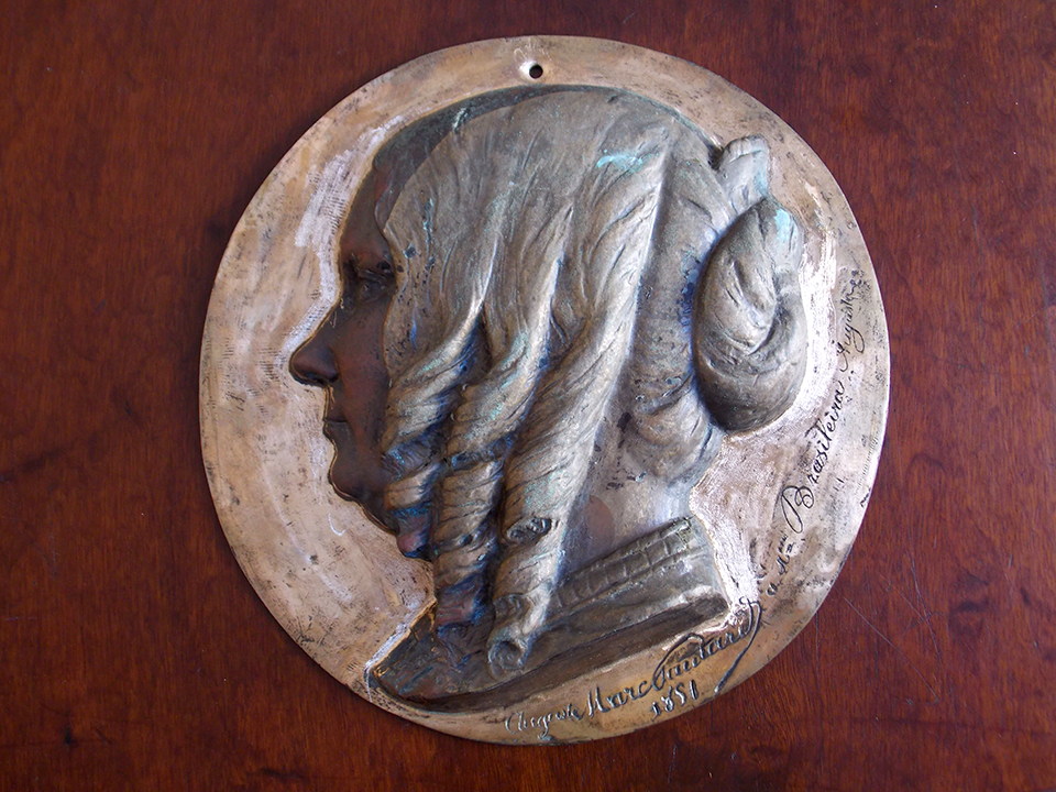 A small bronze medallion depicting Floresta in profile. She faces to the left. Her signature is engraved beneath her portrait.