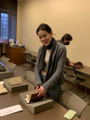 Young woman looks up and smiles as she holds open a small old book that sits on a book rest. 